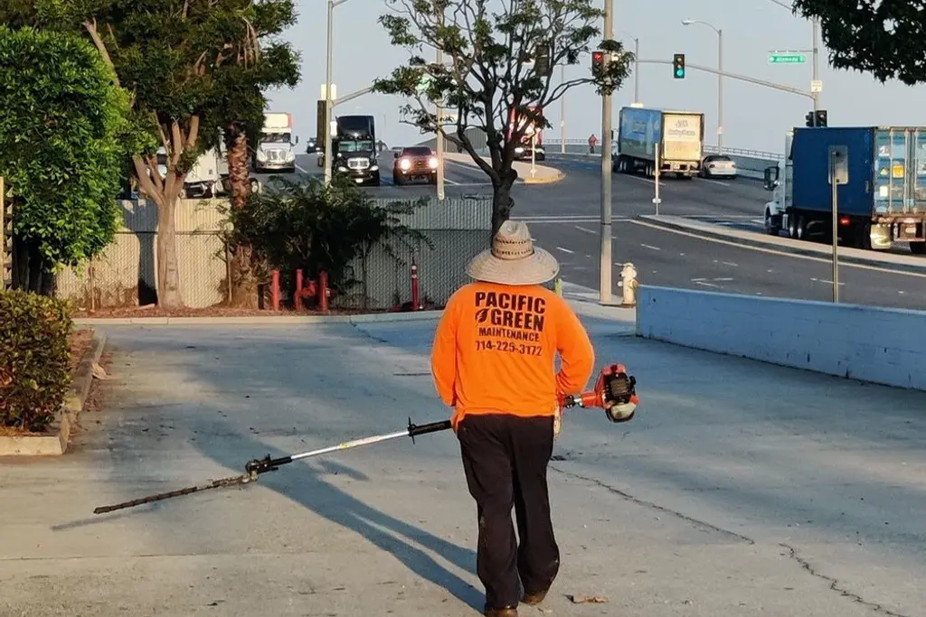 Crew member in a Pacific Green shirt using a blower along a commercial street with delivery trucks behind