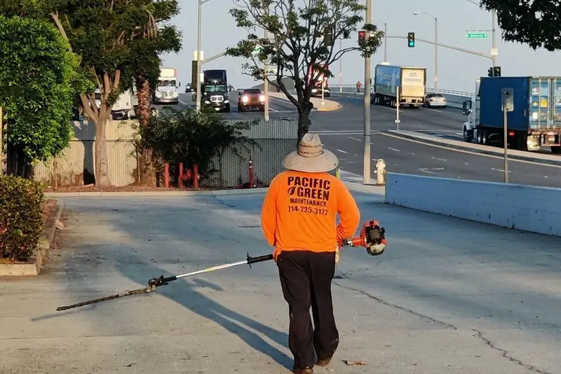 Crew member in a Pacific Green shirt using a blower along a commercial street with delivery trucks behind